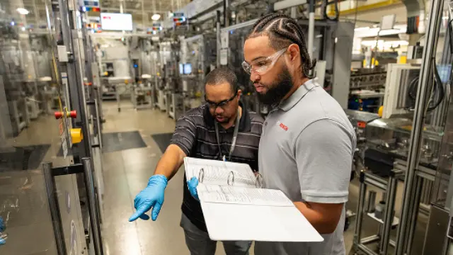 Two people wearing safety glasses and gloves work with a power tool on factory equipment.