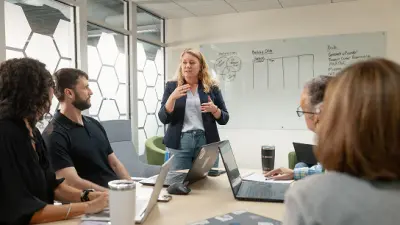 Presenter speaking to colleagues in a meeting room with laptops and a whiteboard.