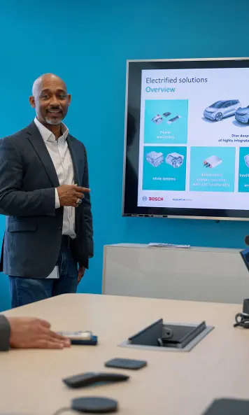 Presenter speaking to colleagues in a meeting room with laptops and a whiteboard.