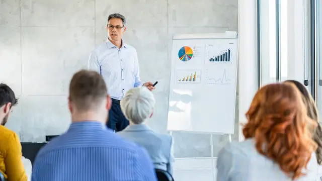 Person stands before seated people and points to flip chart with charts in a meeting room.
