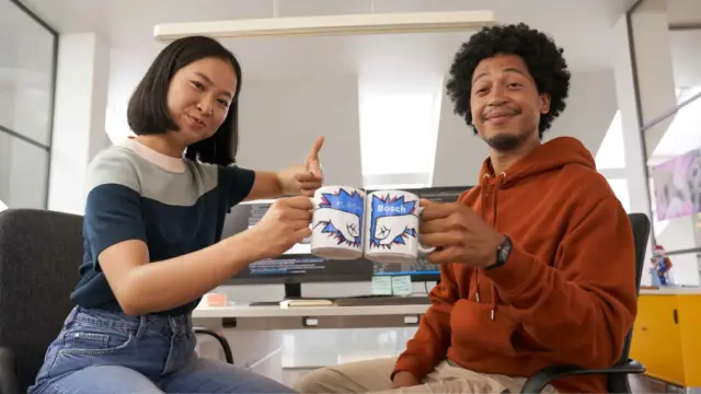 Two people seated at a desk clink branded mugs, with computer screens and notes in background