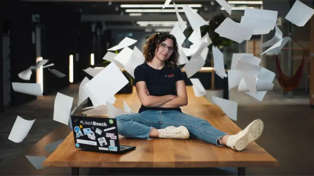 Person sitting cross-legged on a table with arms crossed while sheets of paper float around in an office
