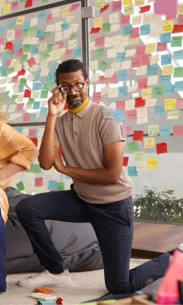 Person kneeling on cushion, adjusting glasses, with wall covered in colorful sticky notes in workspace 