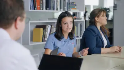 Three people sit at a table with a laptop; shelves in the background.