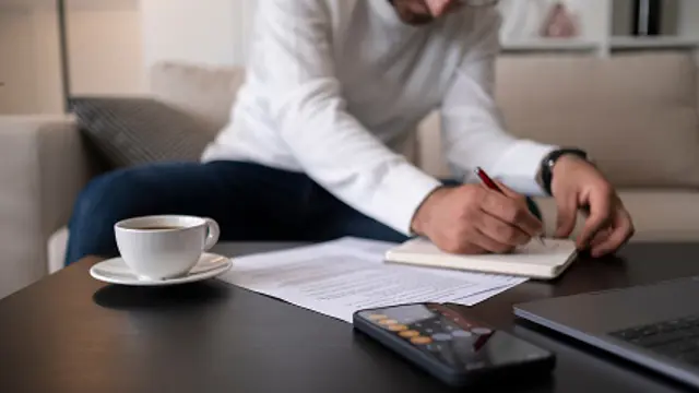 Person writing in a notebook beside documents, a calculator, laptop and coffee.
