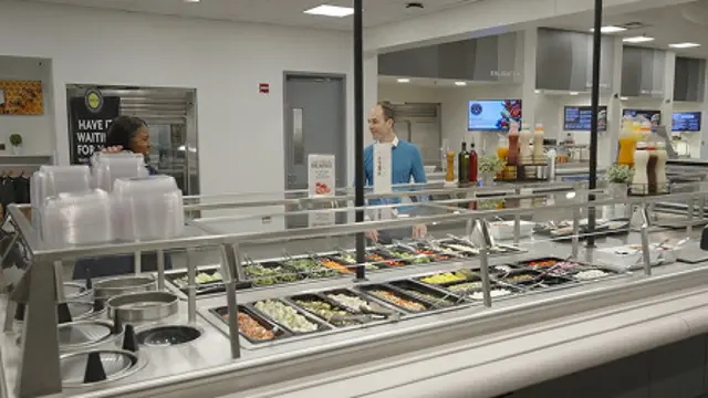Two people standing behind a cafeteria counter with food trays and drink dispensers.