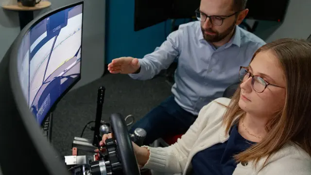 Two people using a driving simulator with a steering wheel and monitor.